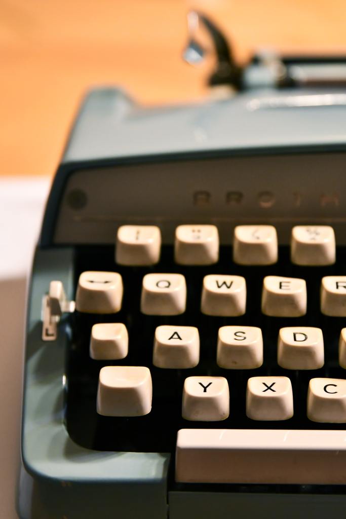 [decorative image] Photo of part of a typewriter on a desk, taken from the front at desk level, showing around 20 keys.  The wall in the background is a bright, citrus colour.